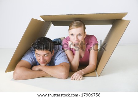 Young smiling couple lying in cardboard box. They're looking at camera. Front view.