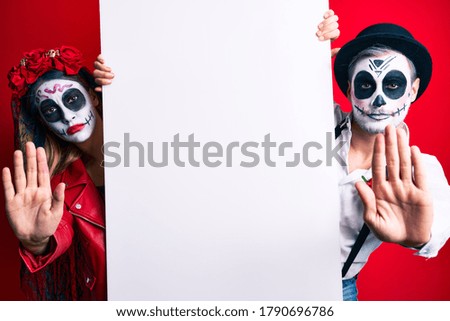 Couple wearing day of the dead costume holding blank empty banner with open hand doing stop sign with serious and confident expression, defense gesture 