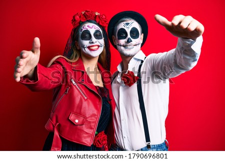 Couple wearing day of the dead costume over red looking at the camera smiling with open arms for hug. cheerful expression embracing happiness. 