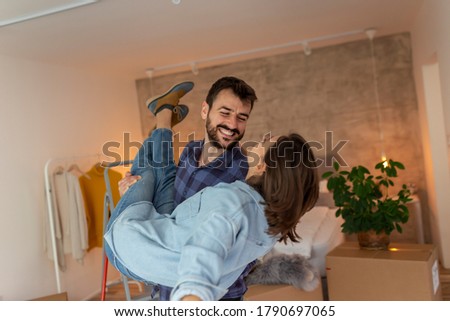 Beautiful young newly married couple moving in together, having fun while unpacking cardboard boxes with their belongings, husband carrying wife in their new home Royalty-Free Stock Photo #1790697065