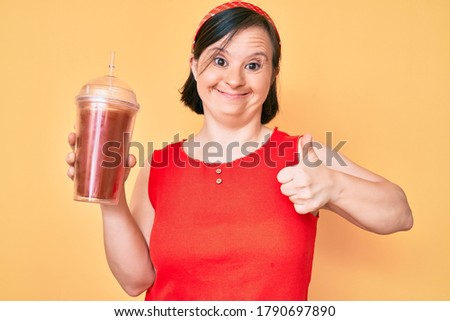 Brunette woman with down syndrome holding glass of smoothie smiling happy and positive, thumb up doing excellent and approval sign 