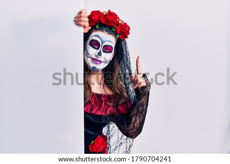 Young woman wearing day of the dead custome holding blank empty banner pointing finger up with successful idea. exited and happy. number one. 
