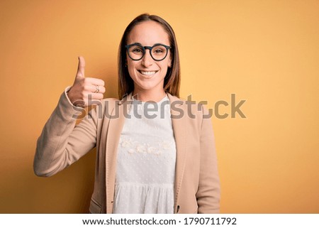 Young beautiful businesswoman wearing jacket and glasses over isolated yellow background doing happy thumbs up gesture with hand. Approving expression looking at the camera showing success.