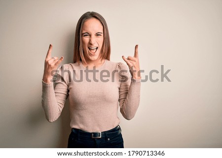Young beautiful woman wearing casual sweater standing over isolated white background shouting with crazy expression doing rock symbol with hands up. Music star. Heavy concept.