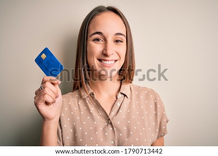 Young beautiful customer woman holding credit card to do payment over white background with a happy face standing and smiling with a confident smile showing teeth