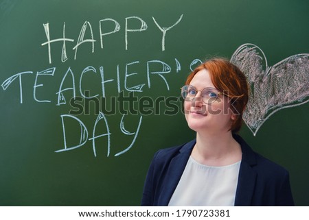 Happy female teacher smiles at the blackboard. Teacher's day greetings and a heart drawn in chalk on a green school chalkboard