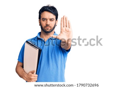 Young hispanic man holding notebook with open hand doing stop sign with serious and confident expression, defense gesture 