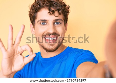 Young caucasian man with curly hair wearing casual clothes taking a selfie doing ok sign with fingers, smiling friendly gesturing excellent symbol 