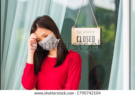 Asian young woman wear face mask protect she sad notice sign wood board label "SORRY WE ARE CLOSED PLEASE COME BACK AGAIN" hanging through glass door front business shop, coronavirus pandemic disease