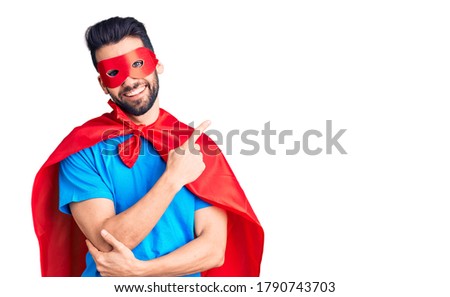 Young handsome man with beard wearing super hero costume with a big smile on face, pointing with hand and finger to the side looking at the camera. 