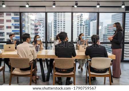 Group of employees sitting at a meeting, wearing a mask, having a leader standing at the head of the table in the meeting, introducing new normal work. Royalty-Free Stock Photo #1790759567