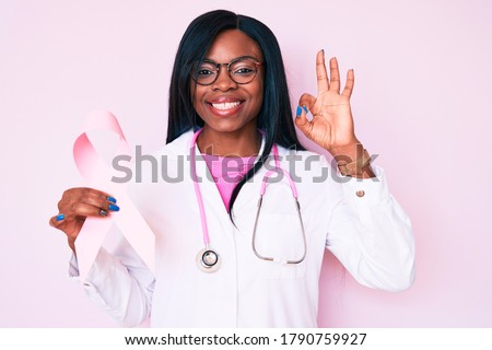 Young african american woman wearing doctor stethoscope holding pink cancer ribbon doing ok sign with fingers, smiling friendly gesturing excellent symbol 