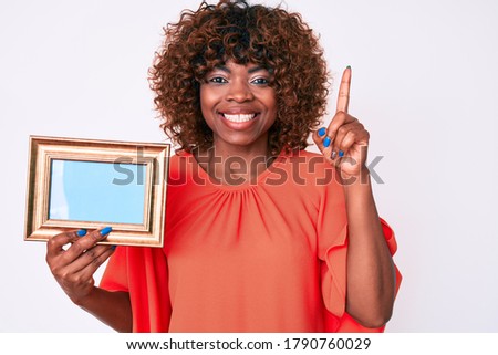 Young african american woman holding empty frame smiling with an idea or question pointing finger with happy face, number one 