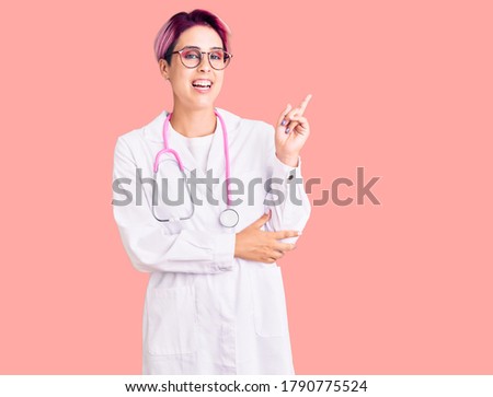Young beautiful woman with pink hair wearing doctor uniform with a big smile on face, pointing with hand and finger to the side looking at the camera. 