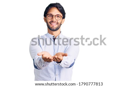Handsome hispanic man wearing business shirt and glasses smiling with hands palms together receiving or giving gesture. hold and protection 