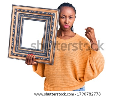 Young african american woman holding empty frame annoyed and frustrated shouting with anger, yelling crazy with anger and hand raised 