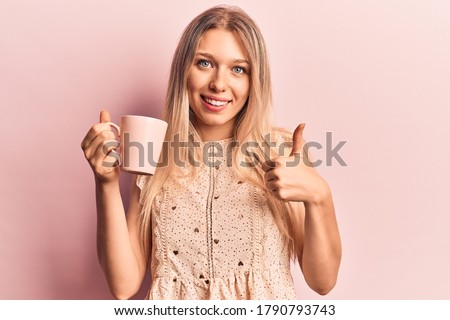 Young beautiful blonde woman holding coffee smiling happy and positive, thumb up doing excellent and approval sign 