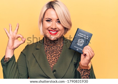 Young beautiful plus size tourist woman holding Italy Itaian passport over yellow background doing ok sign with fingers, smiling friendly gesturing excellent symbol