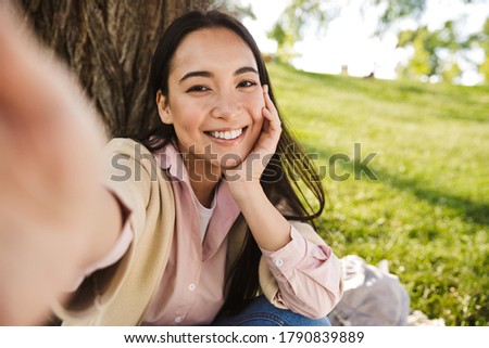 Image of cheerful asian student woman taking selfie photo and smiling while sitting on grass in park