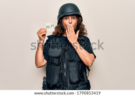 Middle age soldier woman wearing uniform and security helmet holding help reminder paper covering mouth with hand, shocked and afraid for mistake. Surprised expression