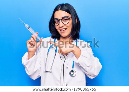Young beautiful latin woman wearing doctor stethoscope holding syringe smiling happy pointing with hand and finger 