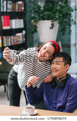 Cheerful young lady smiling and leaning to a positive man by her side while holding a smartphone and taking a selfie