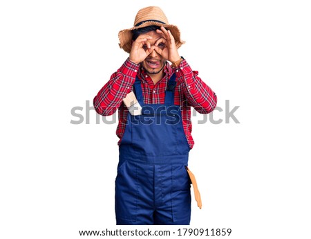 Handsome latin american young man weaing handyman uniform doing ok gesture like binoculars sticking tongue out, eyes looking through fingers. crazy expression. 