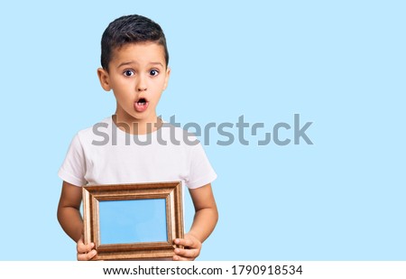 Little cute boy kid holding empty frame scared and amazed with open mouth for surprise, disbelief face 