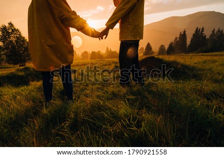 Couple of hikers holding hands standing in the grass in the rain on a background of sunset in the mountains. A couple of tourists were caught in the evening rain on a hike in the mountains. Royalty-Free Stock Photo #1790921558