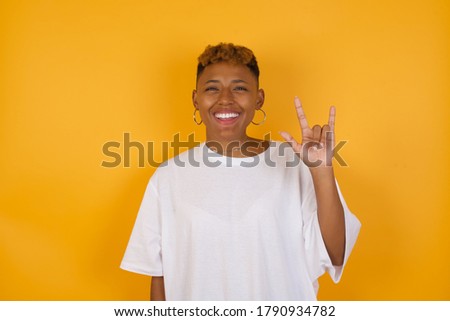 Young african american woman with short hair wearing casual clothes  doing a rock gesture and smiling to the camera. Wearing red shirt standing against white wall. Ready to go to her favorite band 