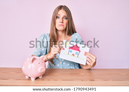 Young blonde girl holding house draw and piggy bank depressed and worry for distress, crying angry and afraid. sad expression. 