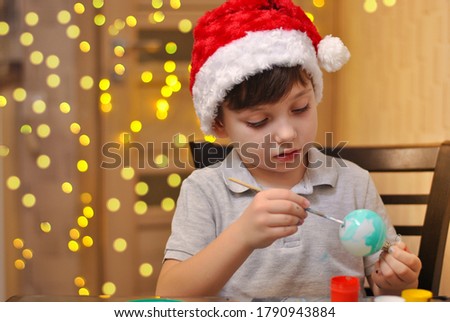 Busy little kid boy paints christmas balls in red and white fluffy xmas hat. Merry holidays children handmade decorations. Bokeh lights background. Children lifestyle