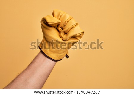 Hand of caucasian young man with gardener glove over isolated yellow background doing protest and revolution gesture, fist expressing force and power