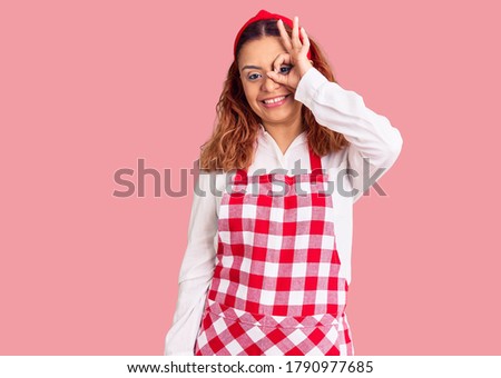 Young latin woman wearing apron smiling happy doing ok sign with hand on eye looking through fingers 