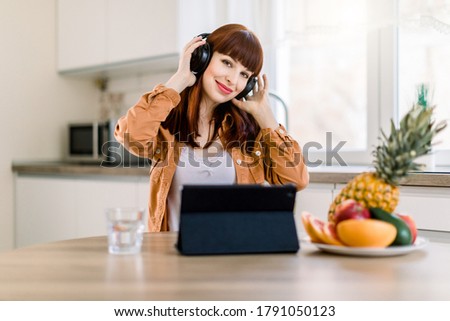 Image of young beautiful smiling woman in headphones, listening to music, while sitting at the table with fresh vegetables and fruits, glass with water and ipad computer in kitchen interior at home