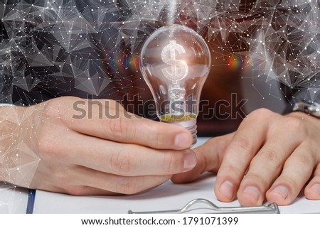 Businessman shows a light bulb with a burning dollar inside on a blurred background.