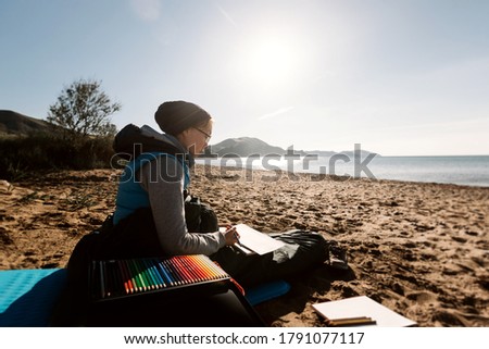 Young woman traveler draws with pencils on the seashore. Cold season.