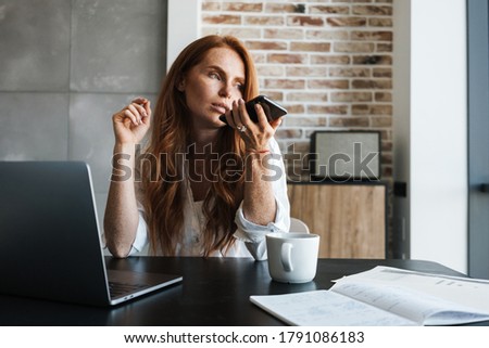 Image of serious businesswoman working with cellphone and laptop while sitting at table in home