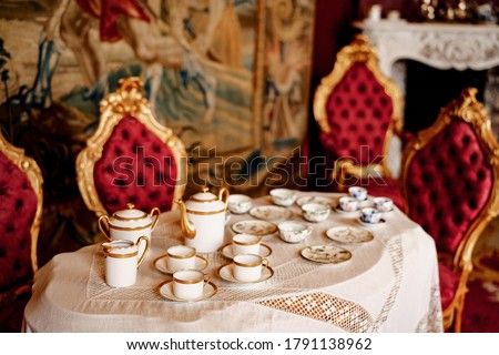 A tea set for four on a table with lace tablecloth and chairs with gold rim and red trim.