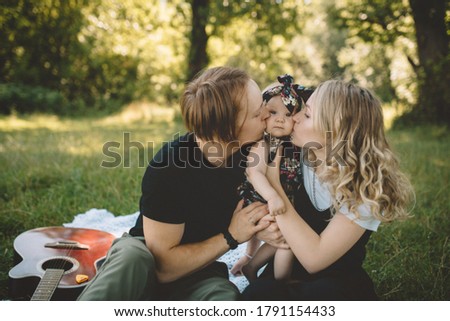 Mommy and daddy kissing their little daughter and in the park. Happy family and childhood. Family picnic with red guitar
