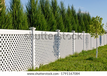 White vinyl fence in a cottage village. Several panels are connected by columns. Tall Thuja bushes behind the fence. Fencing of private property. Royalty-Free Stock Photo #1791167084