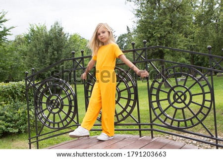 A little girl of six-seven years old with long blond hair in a plain yellow outfit stands and smiles in a summer park on a decorative bridge and poses in full growth.