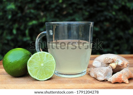 A glass mug filled with lemon and ginger tea. Sliced lemon and ginger roots on the side in a wooden table and green leaves background.
