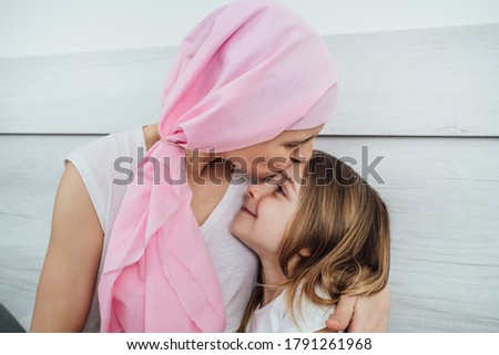 mother with cancer wearing a pink headscarf gives a tender kiss to her beautiful blonde-haired daughter.