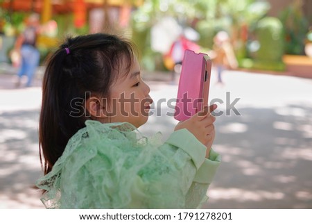 A cute young Asian girl is holding a pink smartphone, trying to take a picture on her vacation. 