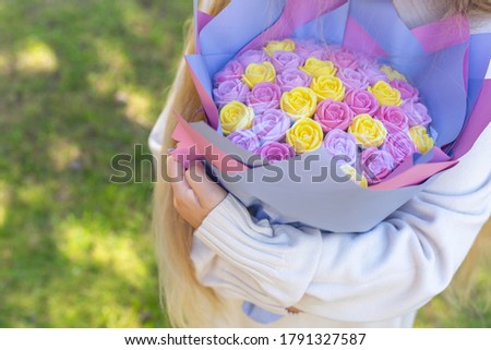 A bouquet of lilac and yellow roses is decorated in multi-colored paper in the hands of a girl on a green natural background. A bouquet of flowers from sweets, soap candies.Birthday gift, mother's day