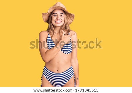 Young beautiful blonde woman wearing bikini and hat cheerful with a smile on face pointing with hand and finger up to the side with happy and natural expression 