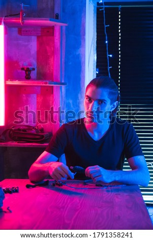 Young man making dream hunter in dark workshop. Artisan at work in futuristic art studio illuminated by neon red and blue lighting. Dreamcatcher creation. Hobby and insomnia concept.