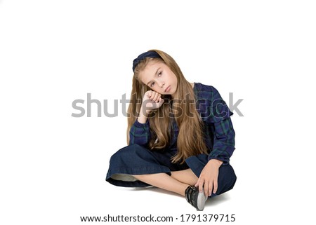 Beautiful schoolgirl girl with long hair sitting cross-legged on floor in blue school clothes on white background. Isolated. Bored, looking at camera. boring education, lack of interest, mute question