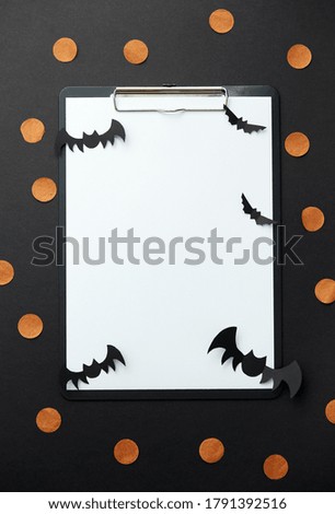 holidays concept - clipboard with sheet of white paper and halloween party decorations on black background